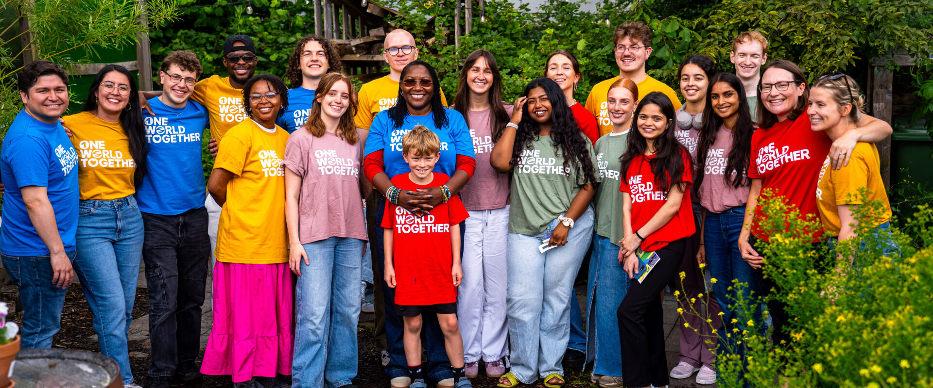 A large group of smiling youth in a vibrant community garden setting, all wearing brightly coloured One World Together tshirts.
