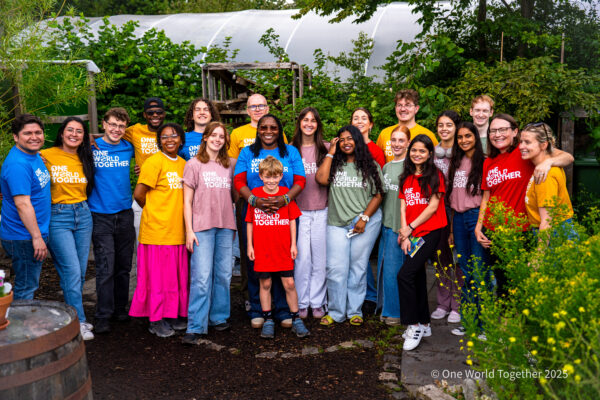 A large group of smiling youth in a vibrant community garden setting, all wearing brightly coloured One World Together tshirts.